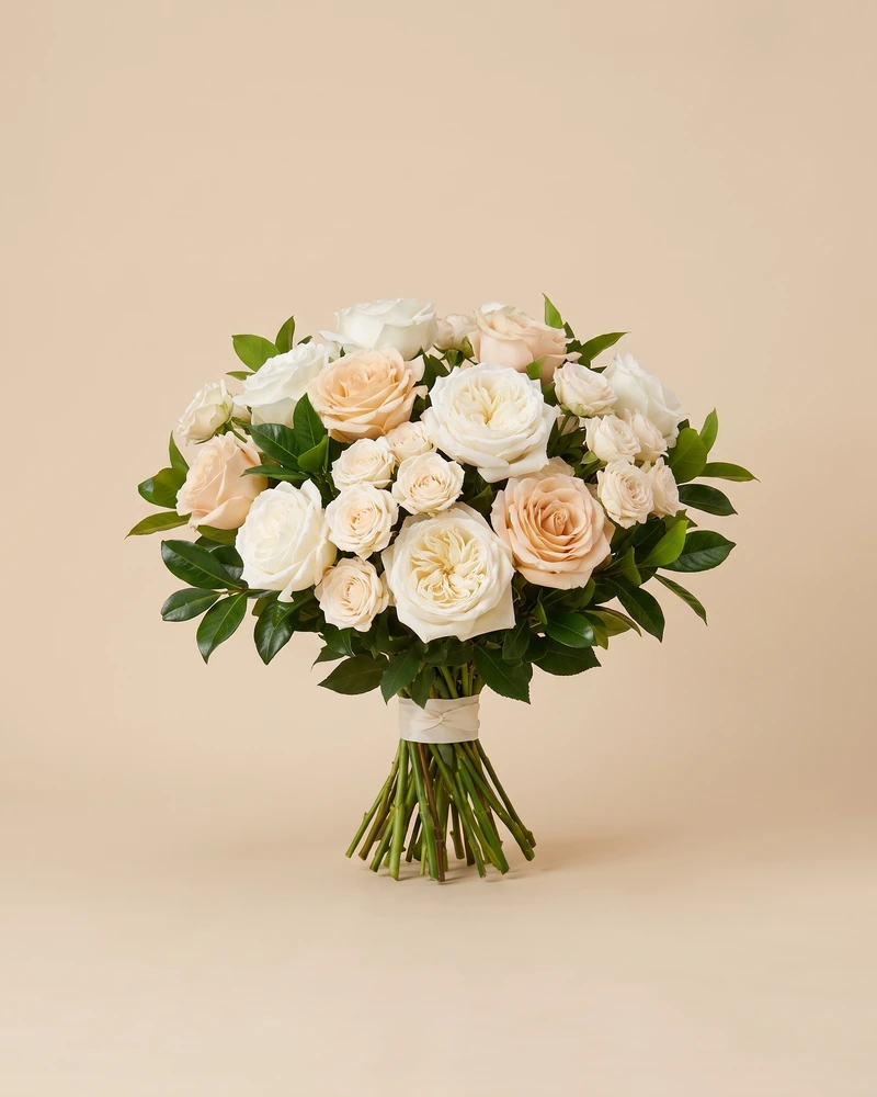 Bouquet rond de roses ivoire, champagne et pêche dans un feuillage vert, tiges liées d’un ruban ivoire.