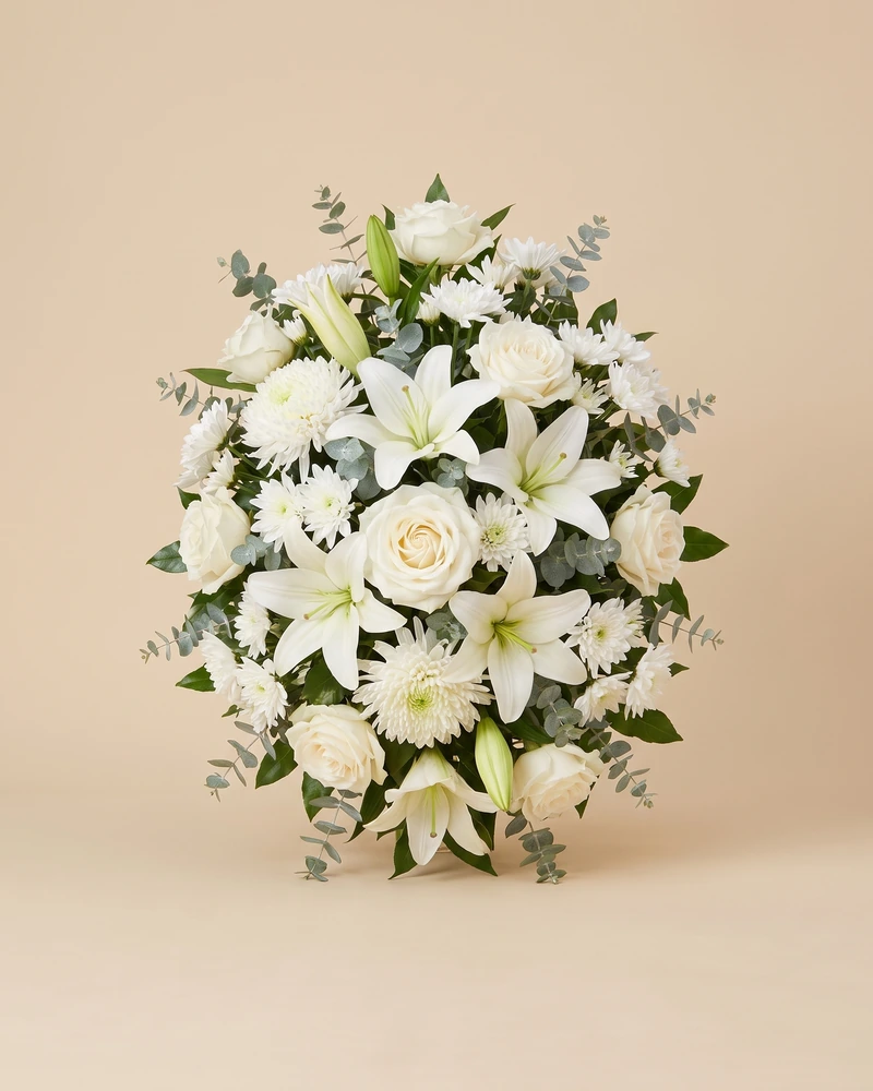 Composition de deuil ovale et symétrique&nbsp;: lys, roses et chrysanthèmes blancs, branches d’eucalyptus.